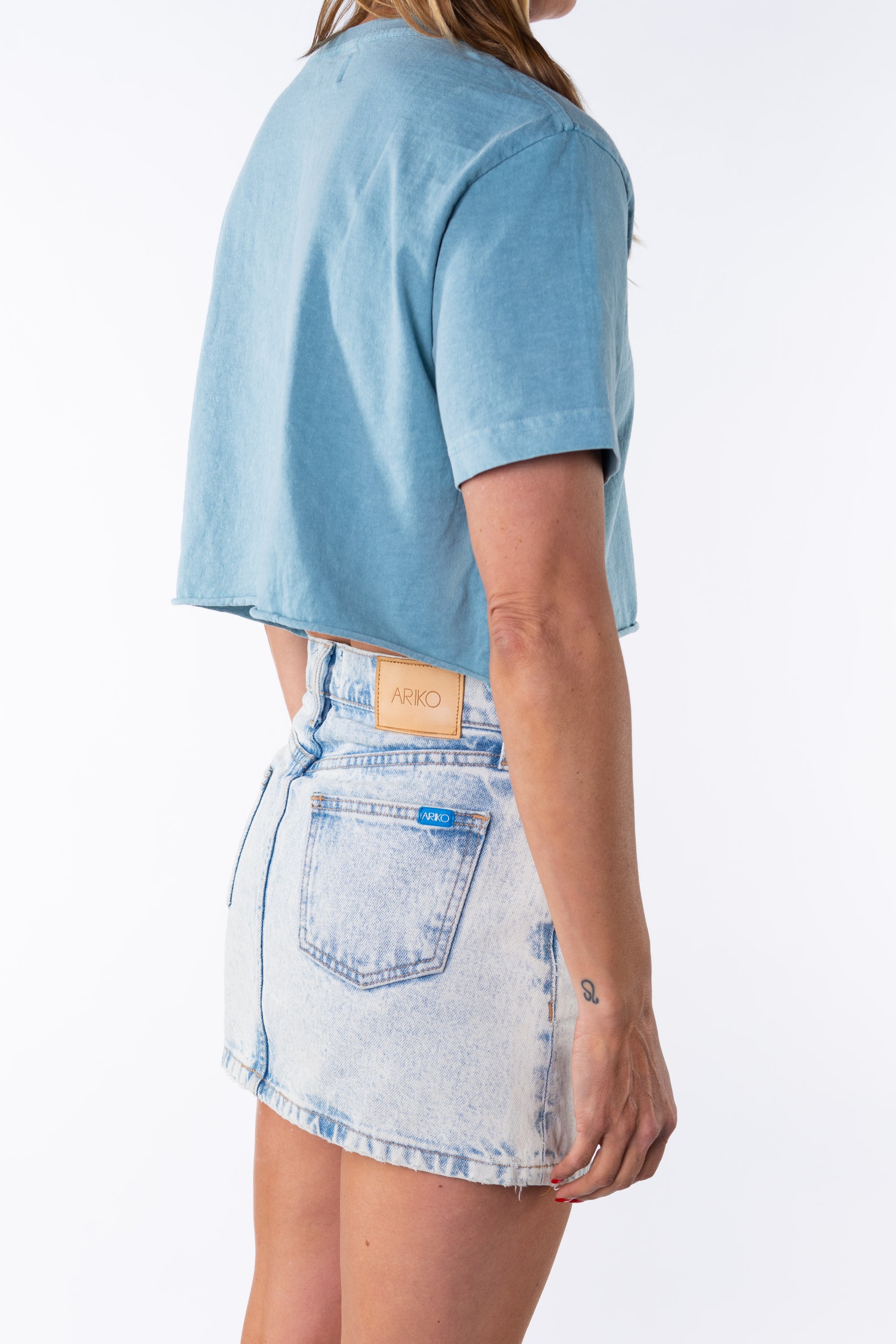 Side back view of a woman wearing a turquoise beach crop t-shirt with a denim mini skirt on a white background. 