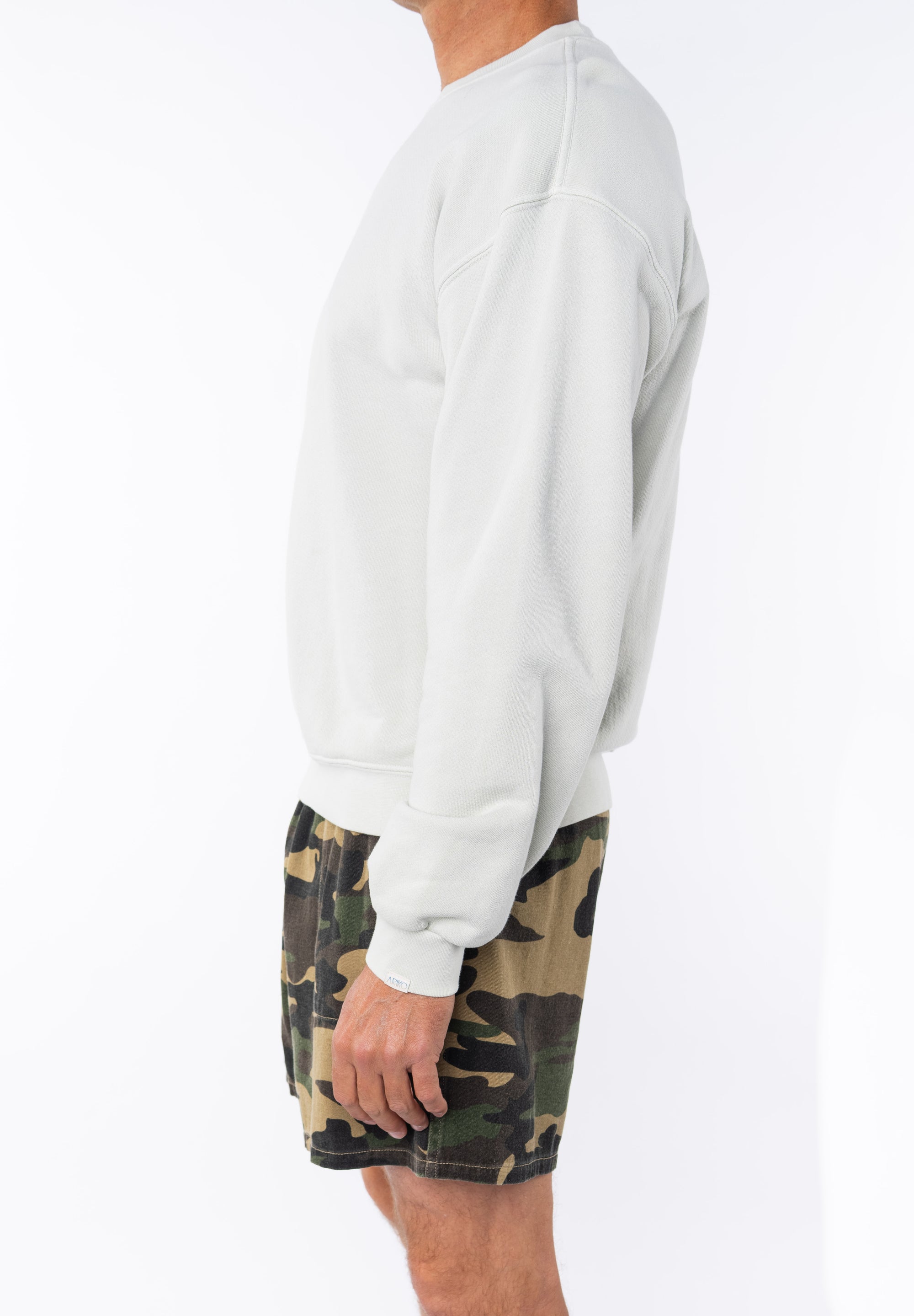 side view of a person wearing a beige sweatshirt and camouflage day shorts on a white background