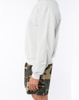 side view of a person wearing a beige sweatshirt and camouflage day shorts on a white background