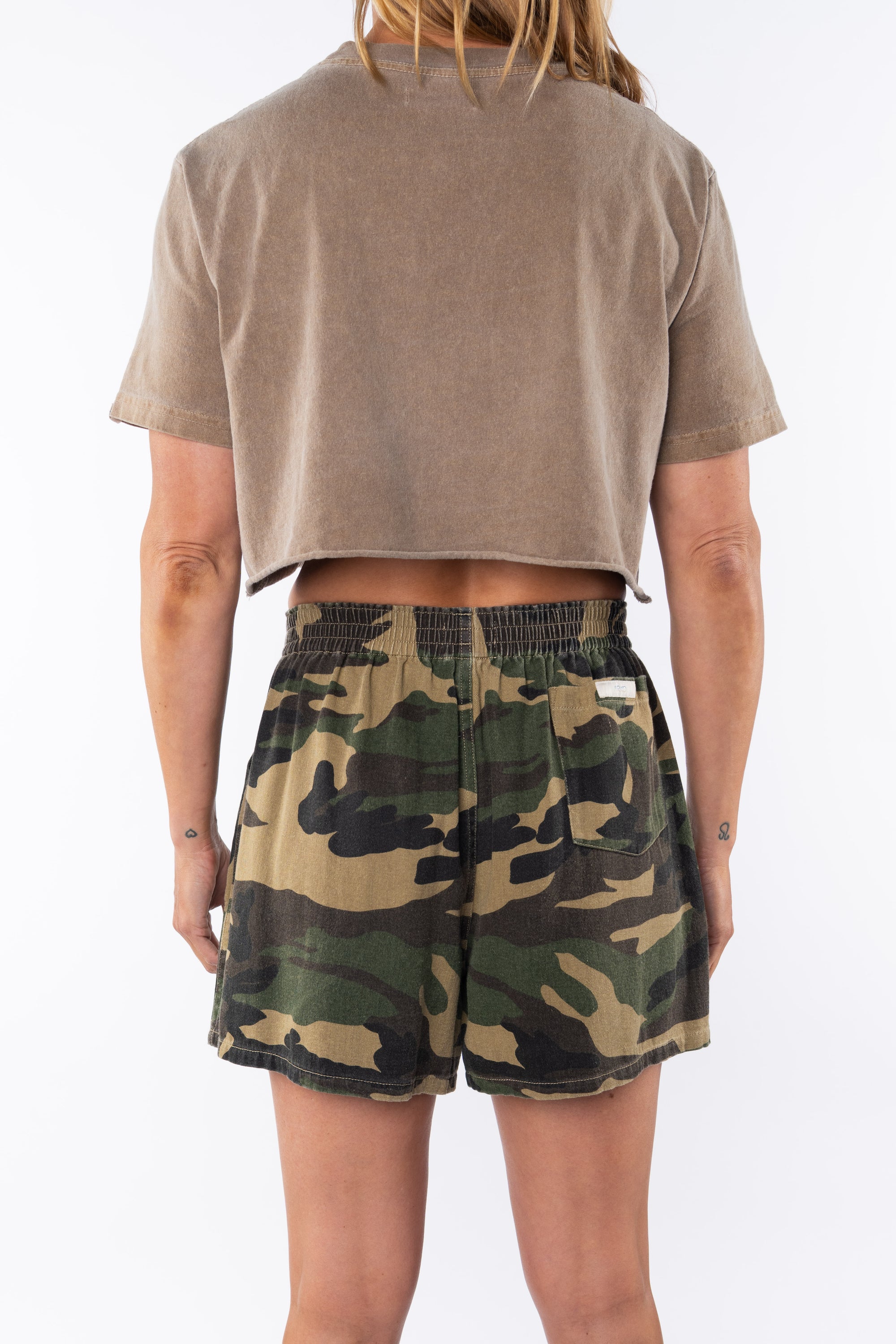 Back view of a woman wearing a brown beach crop t-shirt and camouflage day shorts on a white background