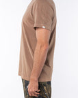 Side view of a person wearing a brown beach t-shirt and real tree camouflage day pants on a white background