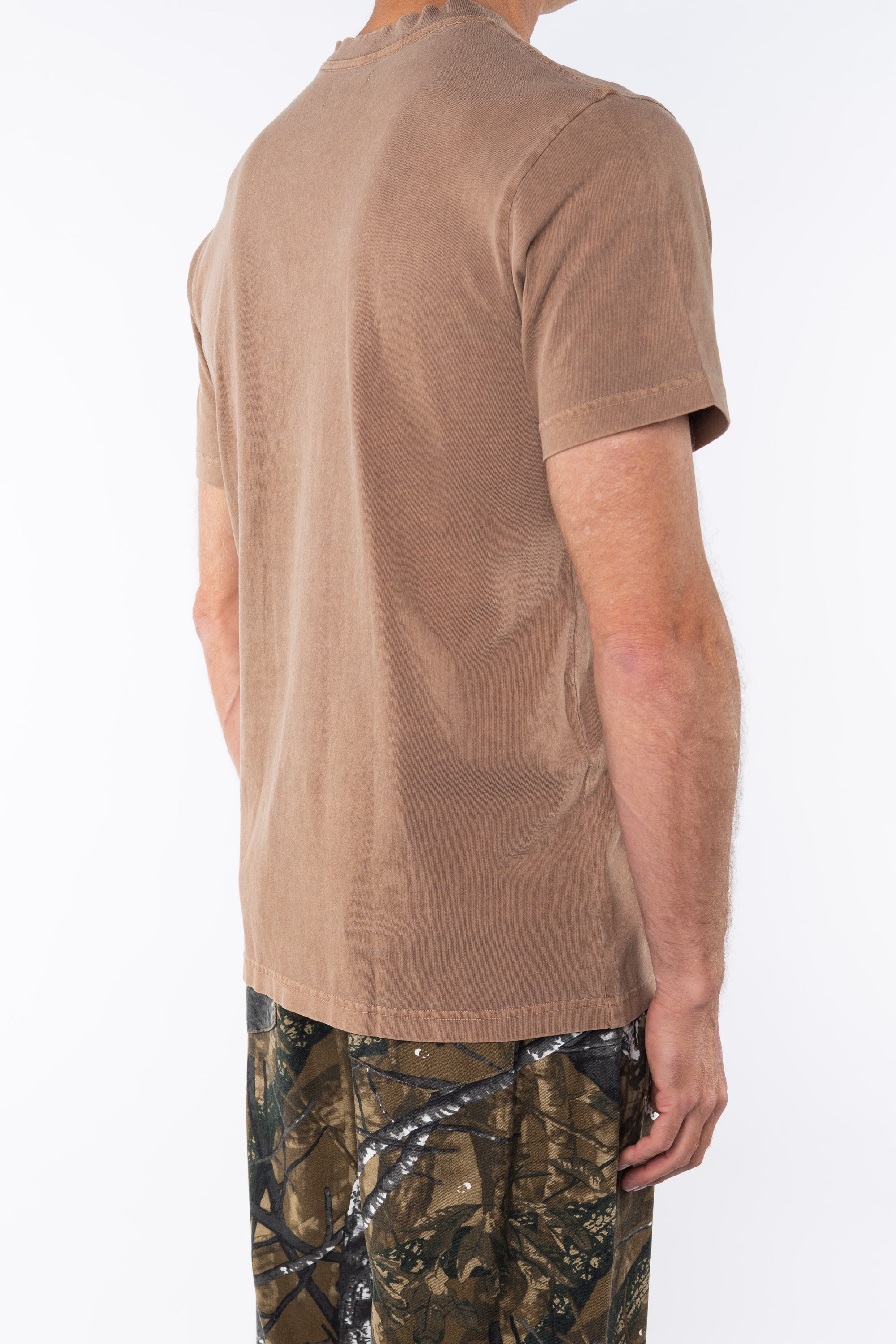 Side view of a person wearing a brown beach t-shirt and real tree camouflage day pants on a white background