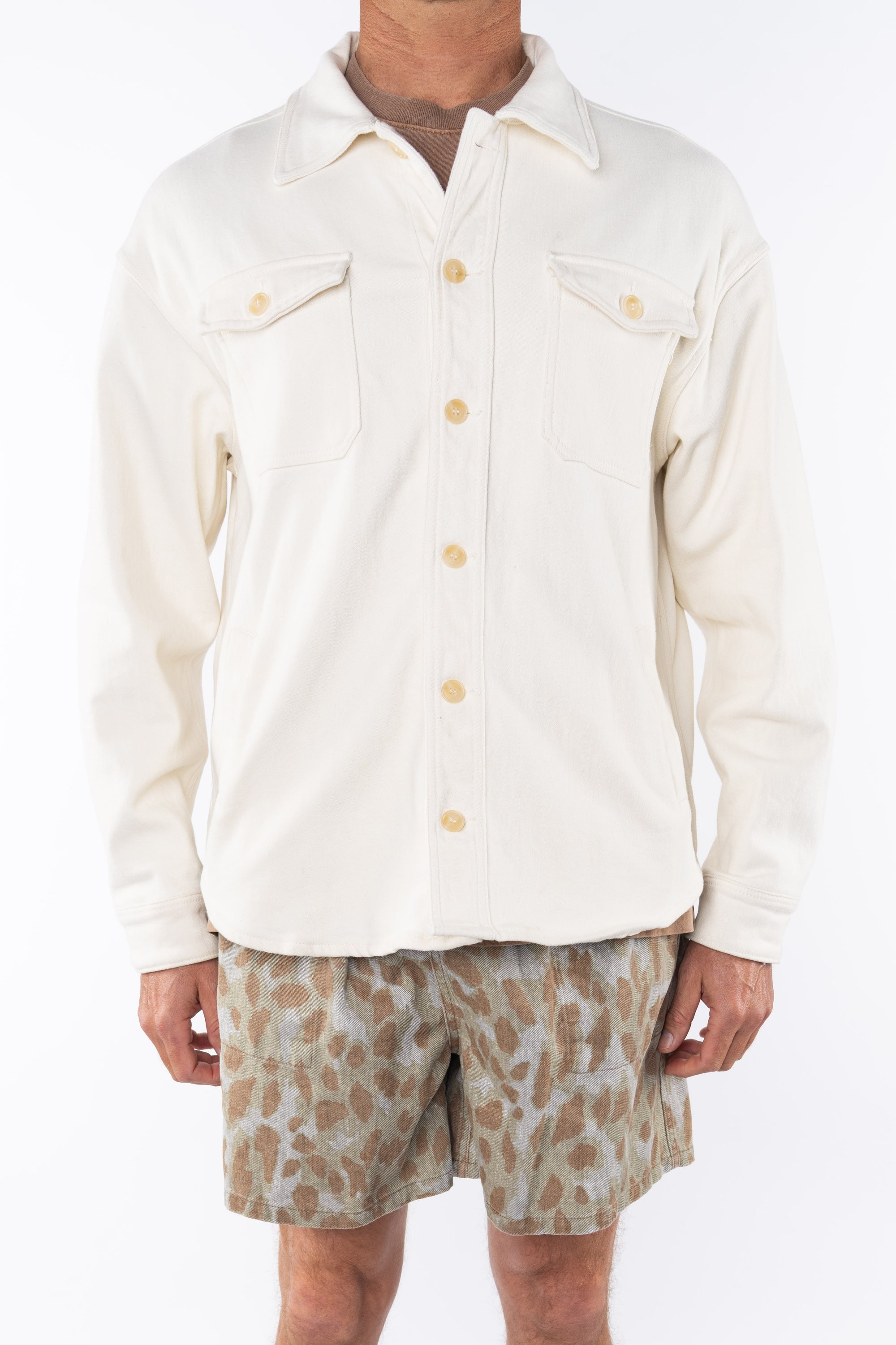 Man wearing a beige bucky shirt jacket buttoned, brown beach t-shirt and leopard camo printed day shorts on a white background