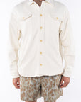 Man wearing a beige bucky shirt jacket buttoned, brown beach t-shirt and leopard camo printed day shorts on a white background
