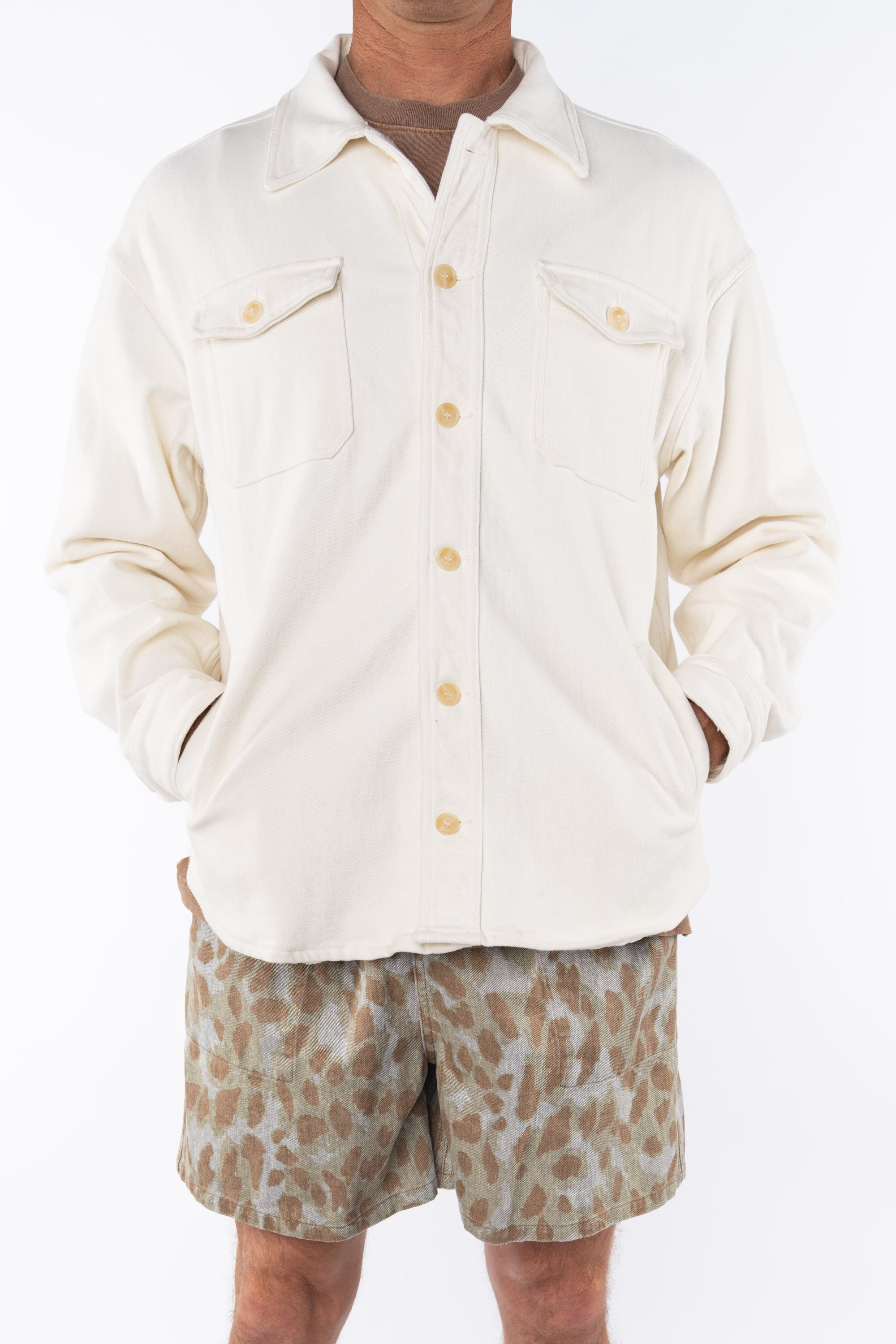 Man wearing a beige bucky shirt jacket buttoned, brown beach t-shirt and leopard camo printed day shorts on a white background