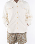 Man wearing a beige bucky shirt jacket buttoned, brown beach t-shirt and leopard camo printed day shorts on a white background