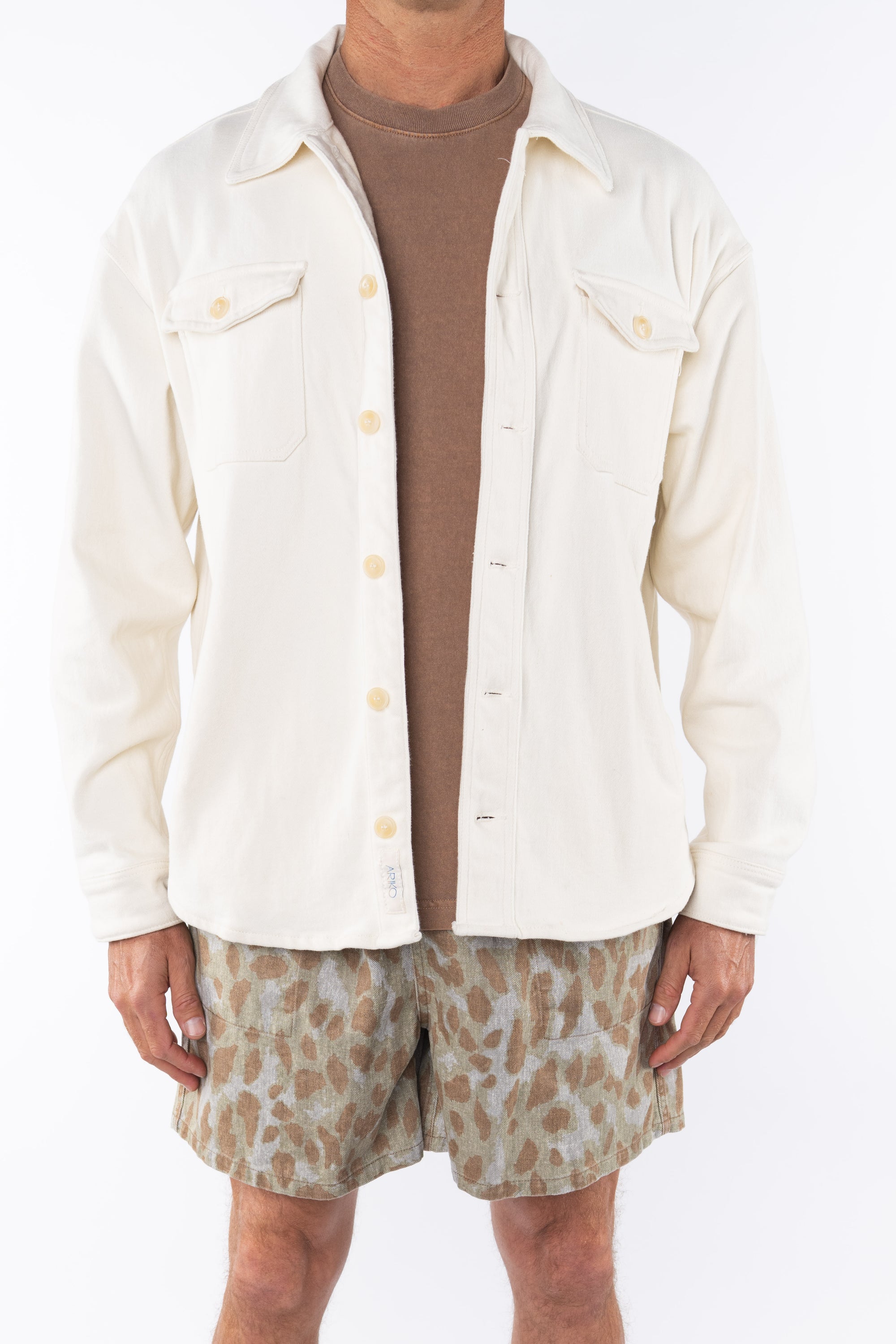 Man wearing a beige bucky shirt jacket, brown beach t-shirt and leopard camo printed day shorts on a white background