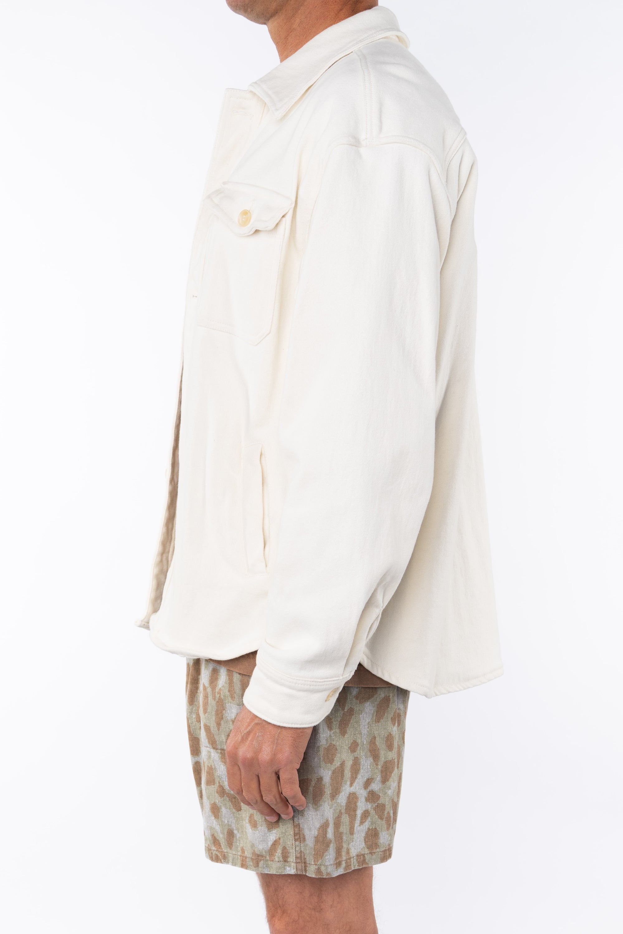 Side view of a man wearing a beige bucky shirt jacket, brown beach t-shirt and leopard camo printed day shorts on a white background