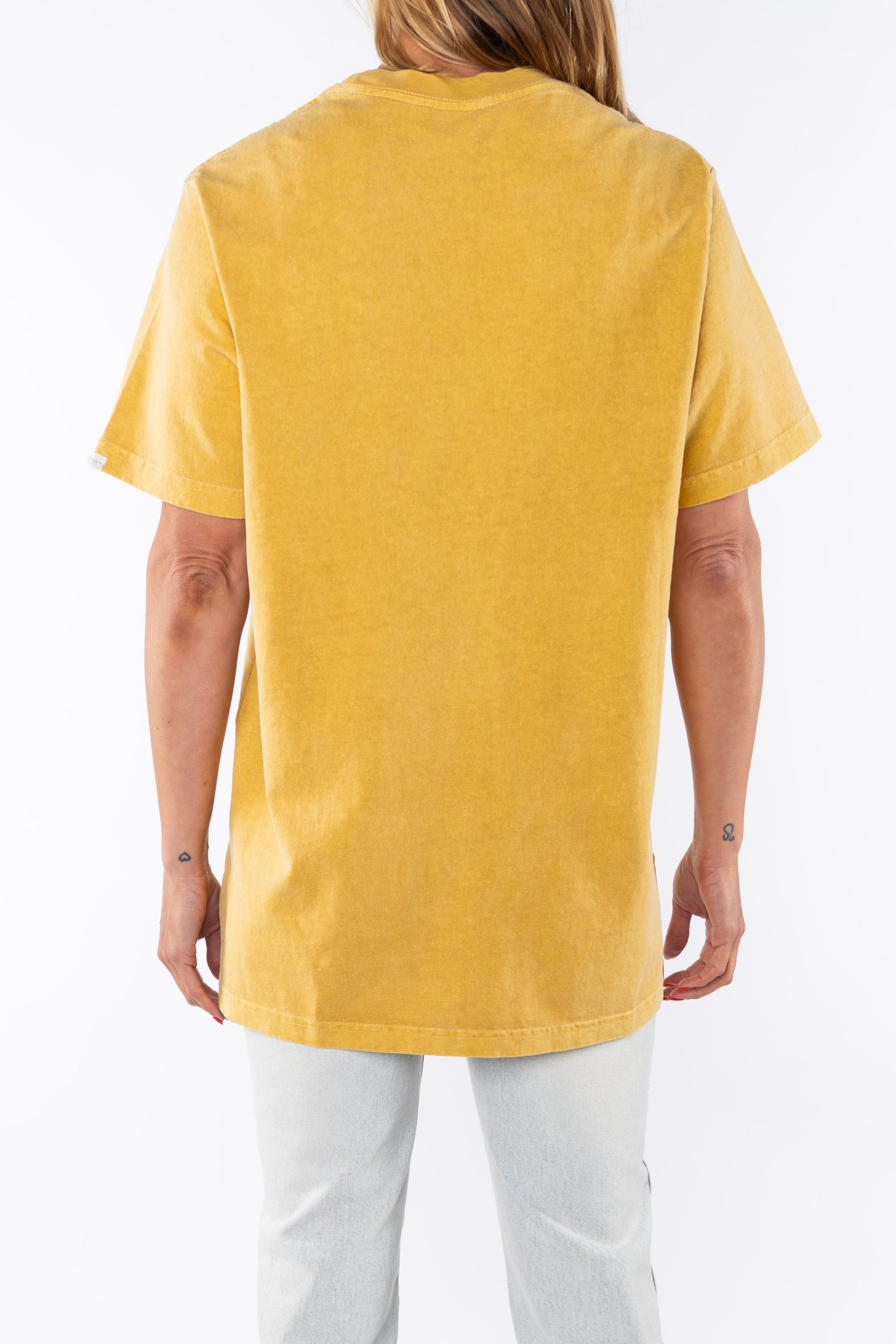 Back view of a woman wearing a sunrise yellow beach t-shirt and light denim jeans on a white background