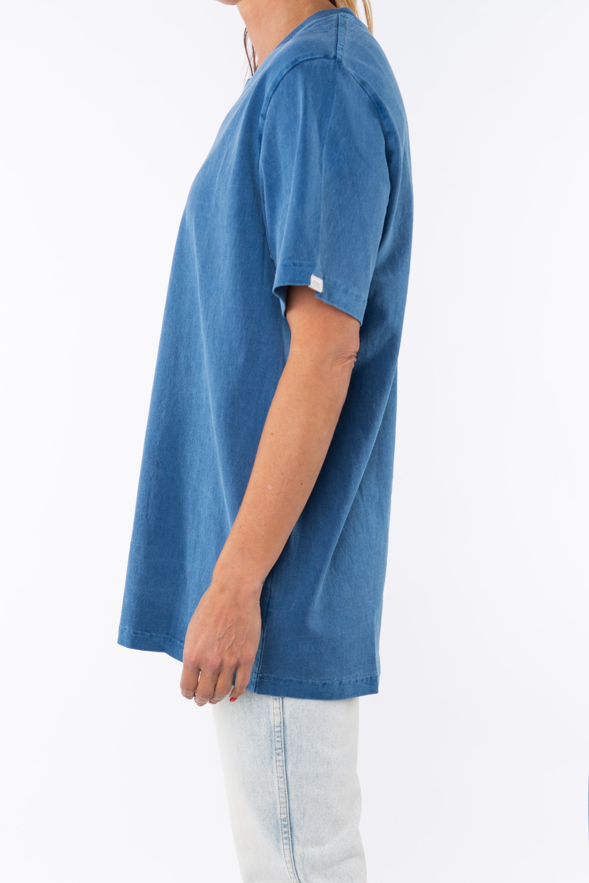Side view of a woman wearing a teal beach t-shirt and light denim jeans on a white background