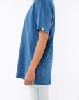 Side view of a woman wearing a teal beach t-shirt and light denim jeans on a white background