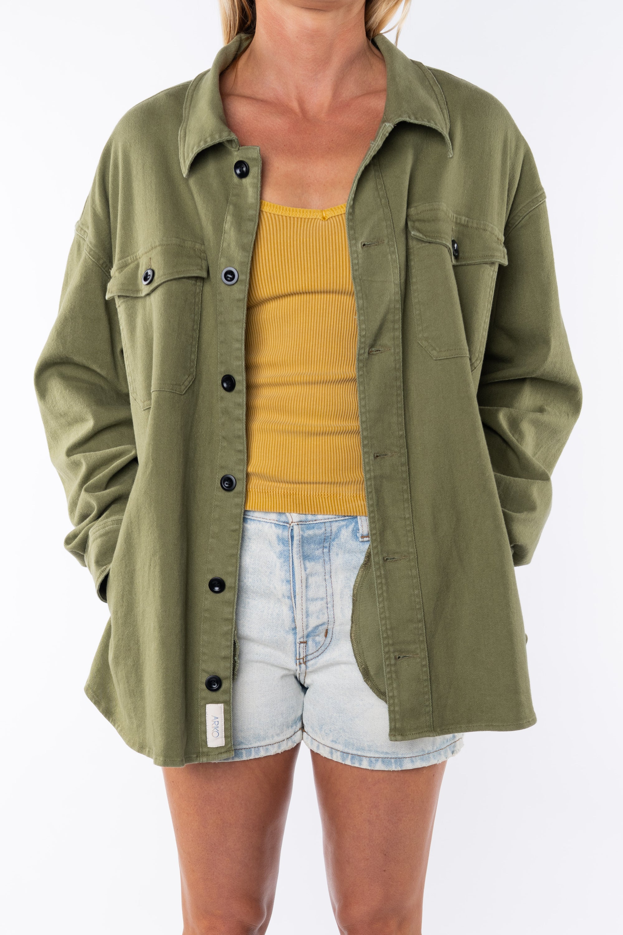 Woman wearing a twill shirt Jacket in army green with a v-neck tank in yellow and denim shorts. 