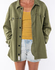 Woman wearing a twill shirt Jacket in army green with a v-neck tank in yellow and denim shorts. 