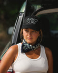 Woman wearing a logo trucker hat with "ARIKO SURF" printed on it. She has a tank top on and a beach bandana. 