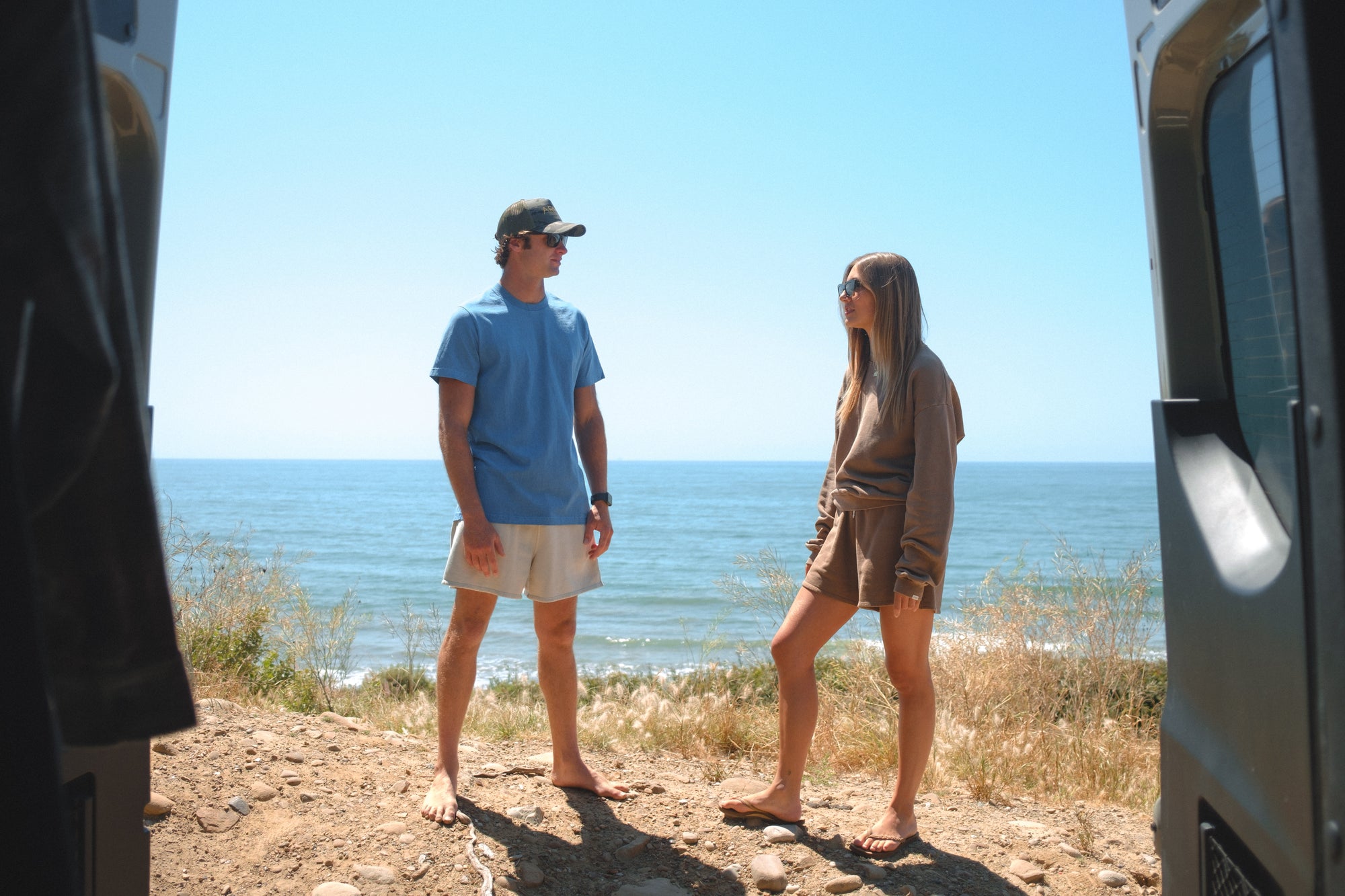 Two people standing on a coastal road with a clear blue sky and ocean view. Man is wearing blue beach t-shirt with beige french terry day shorts. Woman is wearing brown post surf crew sweatshirt and french terry day shorts. 