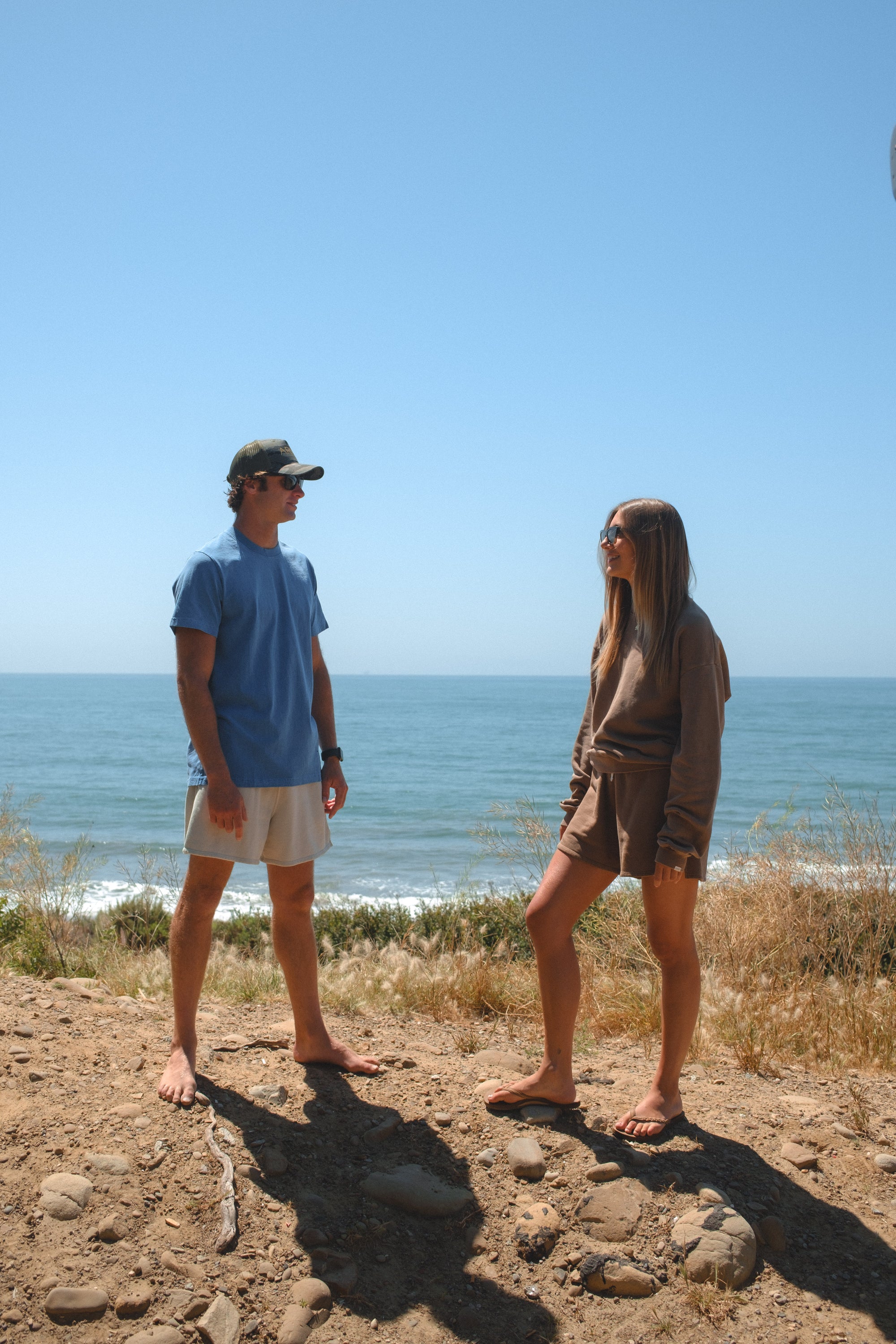 Two people standing on a dirt path by the ocean under a clear blue sky. Man is wearing blue beach t-shirt and beige french terry day shorts and ocean tiger surf trucker hat.