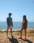Two people standing on a dirt path by the ocean under a clear blue sky. Man is wearing blue beach t-shirt and beige french terry day shorts and ocean tiger surf trucker hat.
