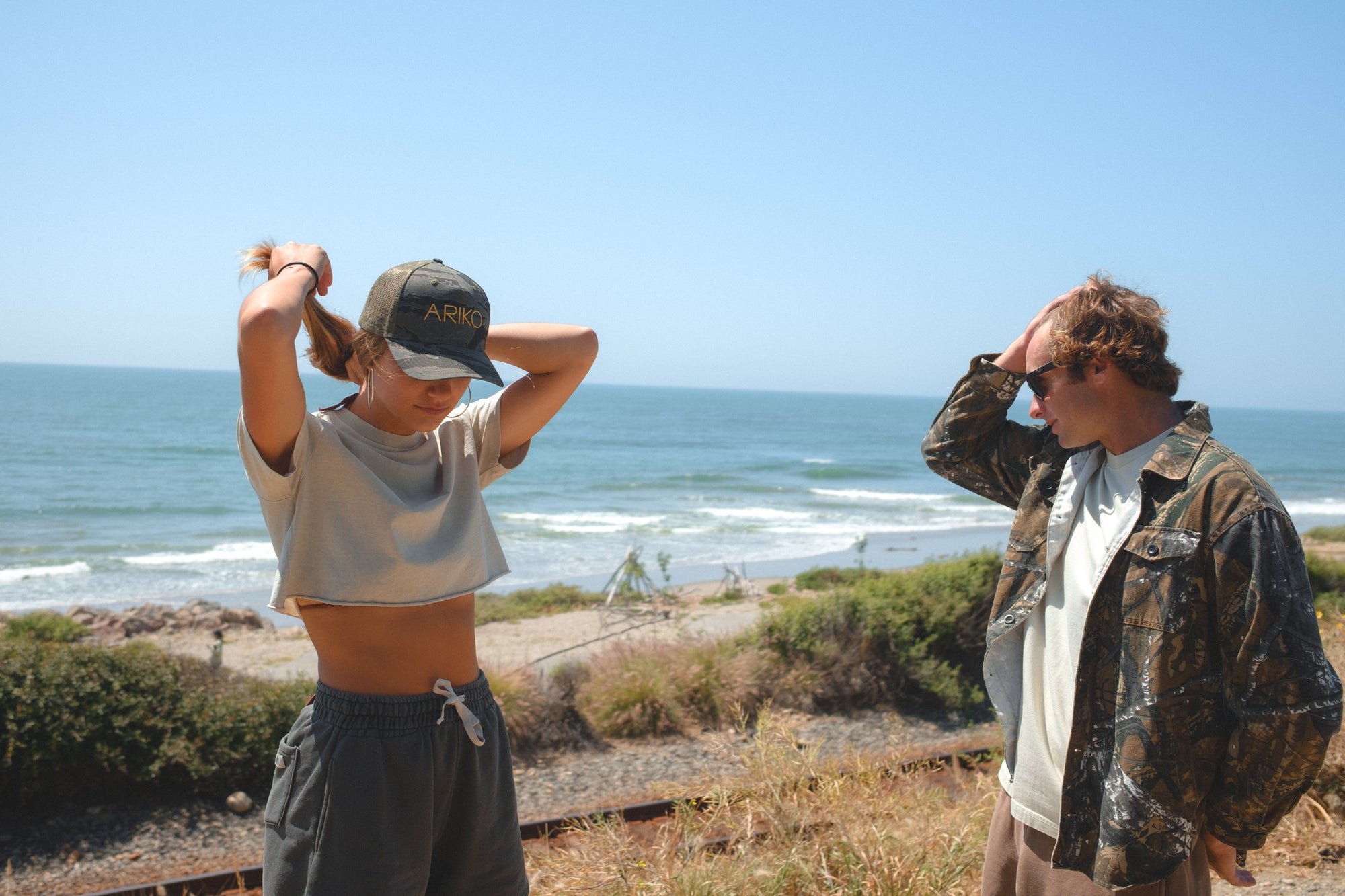Two people standing on a beach with ocean view. She is wearing a beige beach crop t-shirt, dark gray french terry day shorts and an ocean tiger surf trucker hat. He is wearing a real tree camo bucky jacket. 