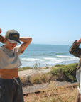 Two people standing on a beach with ocean view. She is wearing a beige beach crop t-shirt, dark gray french terry day shorts and an ocean tiger surf trucker hat. He is wearing a real tree camo bucky jacket. 