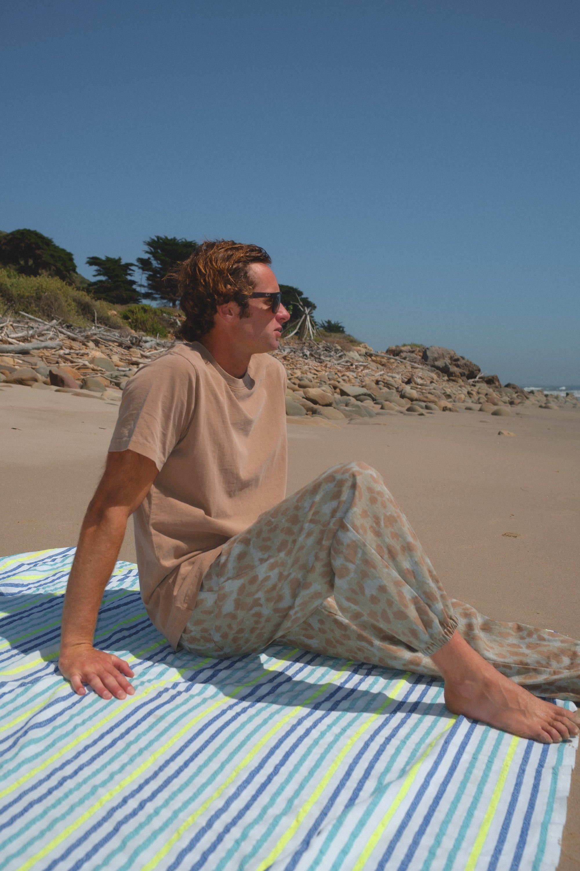 Person sitting on a striped towel at the beach with a clear sky. He is wearing beach t-shirt in brown and leopard camo day pants. 