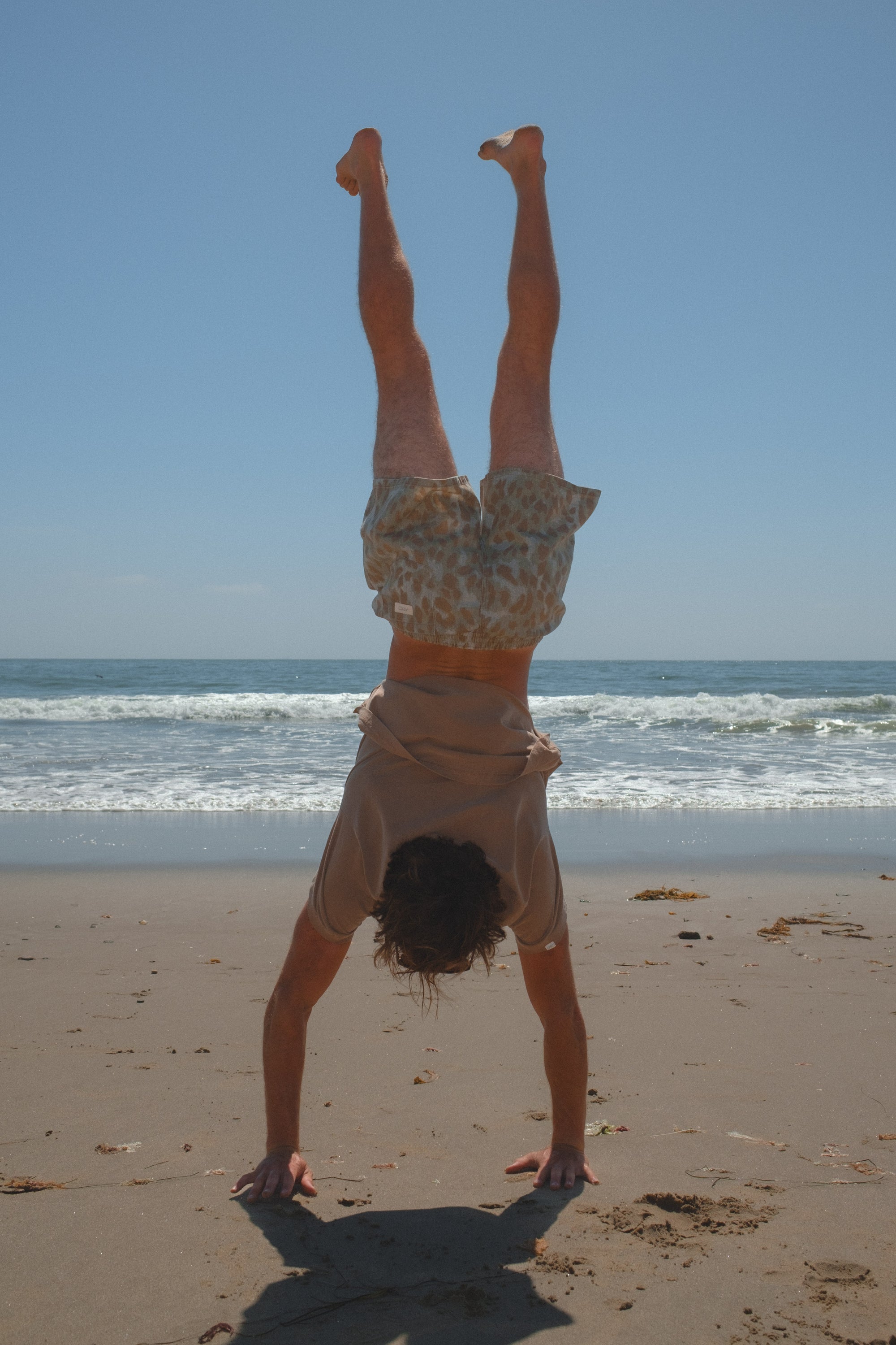 Person doing a handstand on a beach with ocean waves in the background. He is wearing brown beach t-shirt and leopard camo day shorts. 