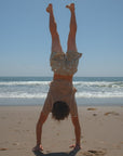 Person doing a handstand on a beach with ocean waves in the background. He is wearing brown beach t-shirt and leopard camo day shorts. 