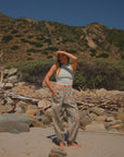 Person standing on a rocky beach with mountains in the background. She is wearing a scallop tank in beige and leopard camo day pants. 