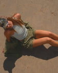 Person lying on a sandy surface wearing an army green shirt, day shorts and beige scallop tank and a real tree surf trucker hat. 