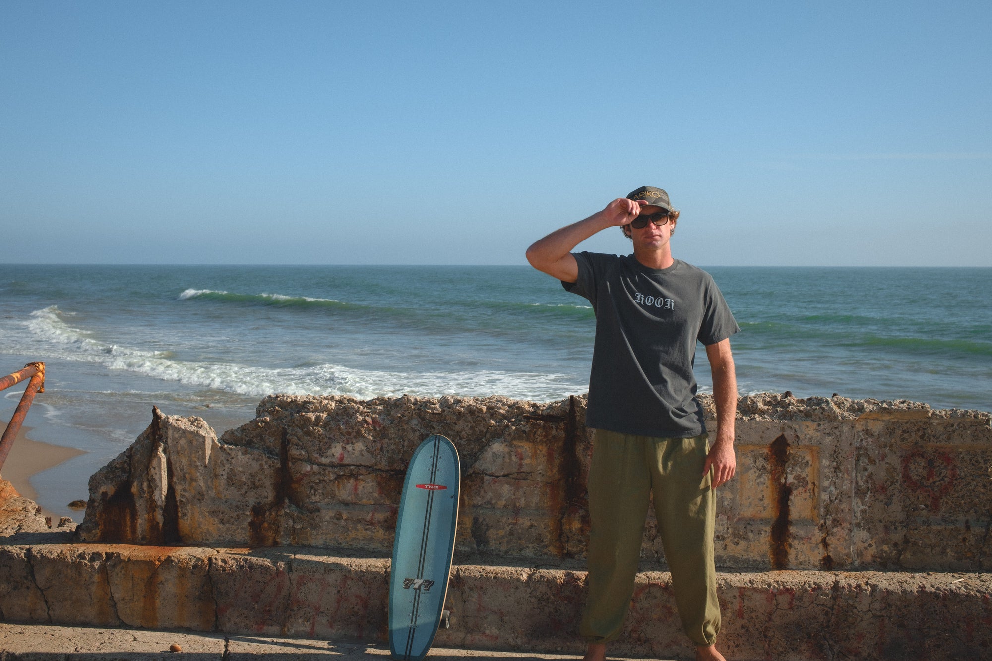 Person standing on a beach with a skateboard, wearing a surf trucker hat in ocean tiger camo, a 'KOOK' graphic t-shirt, and army green day pants.