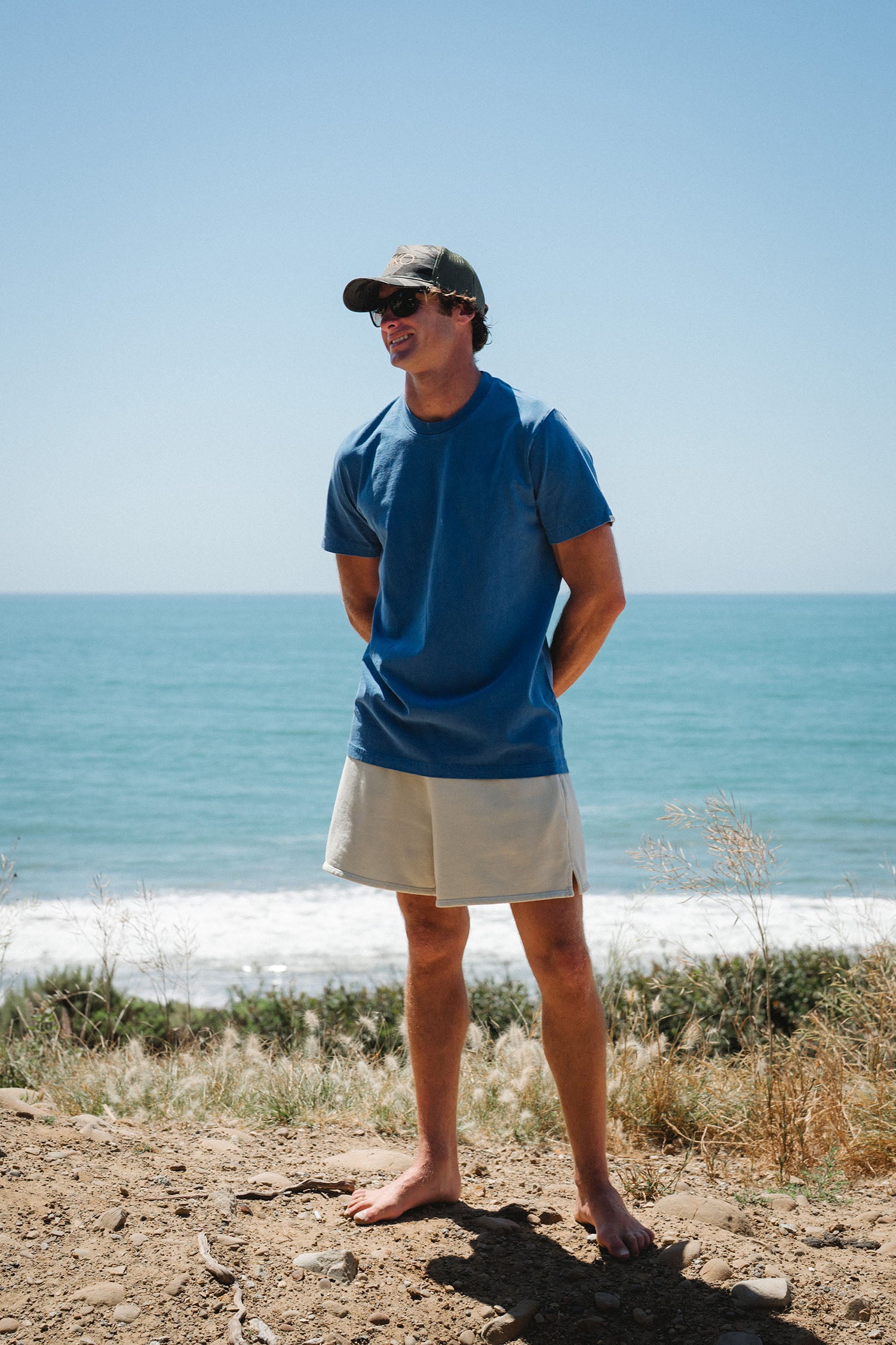Man standing on a beach wearing a blue beach t-shirt and beige french terry day shorts and ocean tiger surf trucker hat.