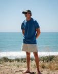 Man standing on a beach wearing a blue beach t-shirt and beige french terry day shorts and ocean tiger surf trucker hat.