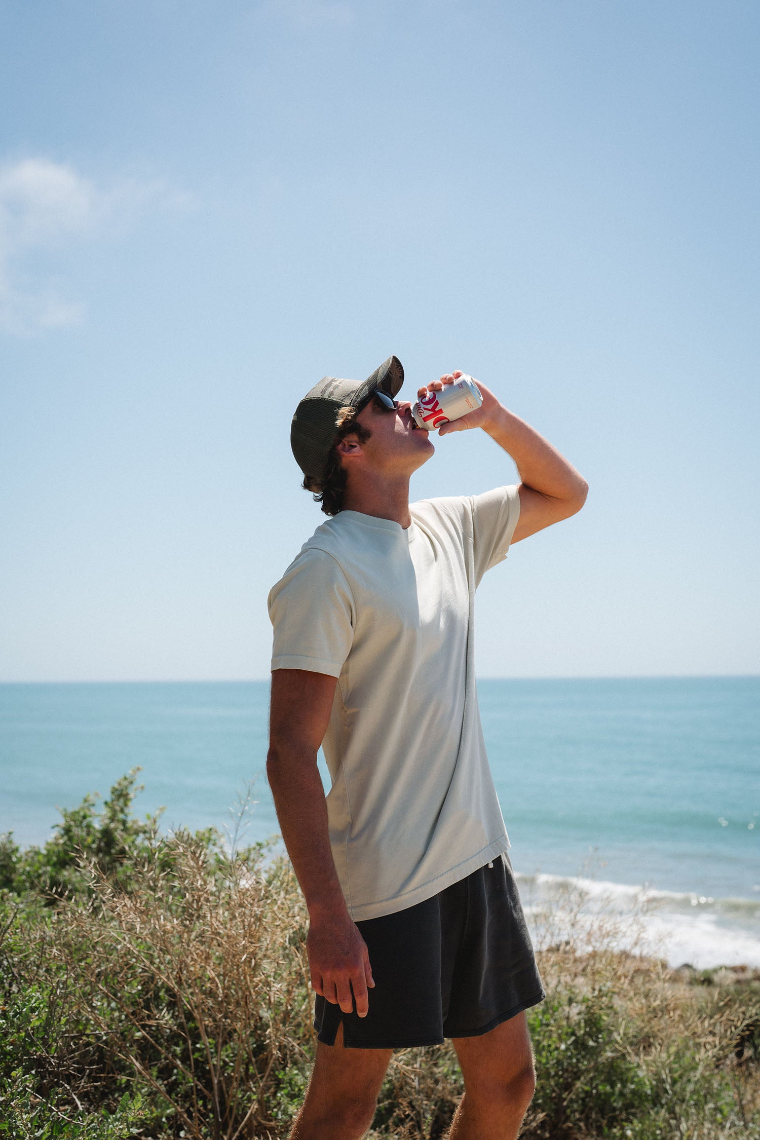Man drinking a soda by the ocean on a clear day. He is wearing a beige beach t-shirt and french terry day shorts in dark gray. 