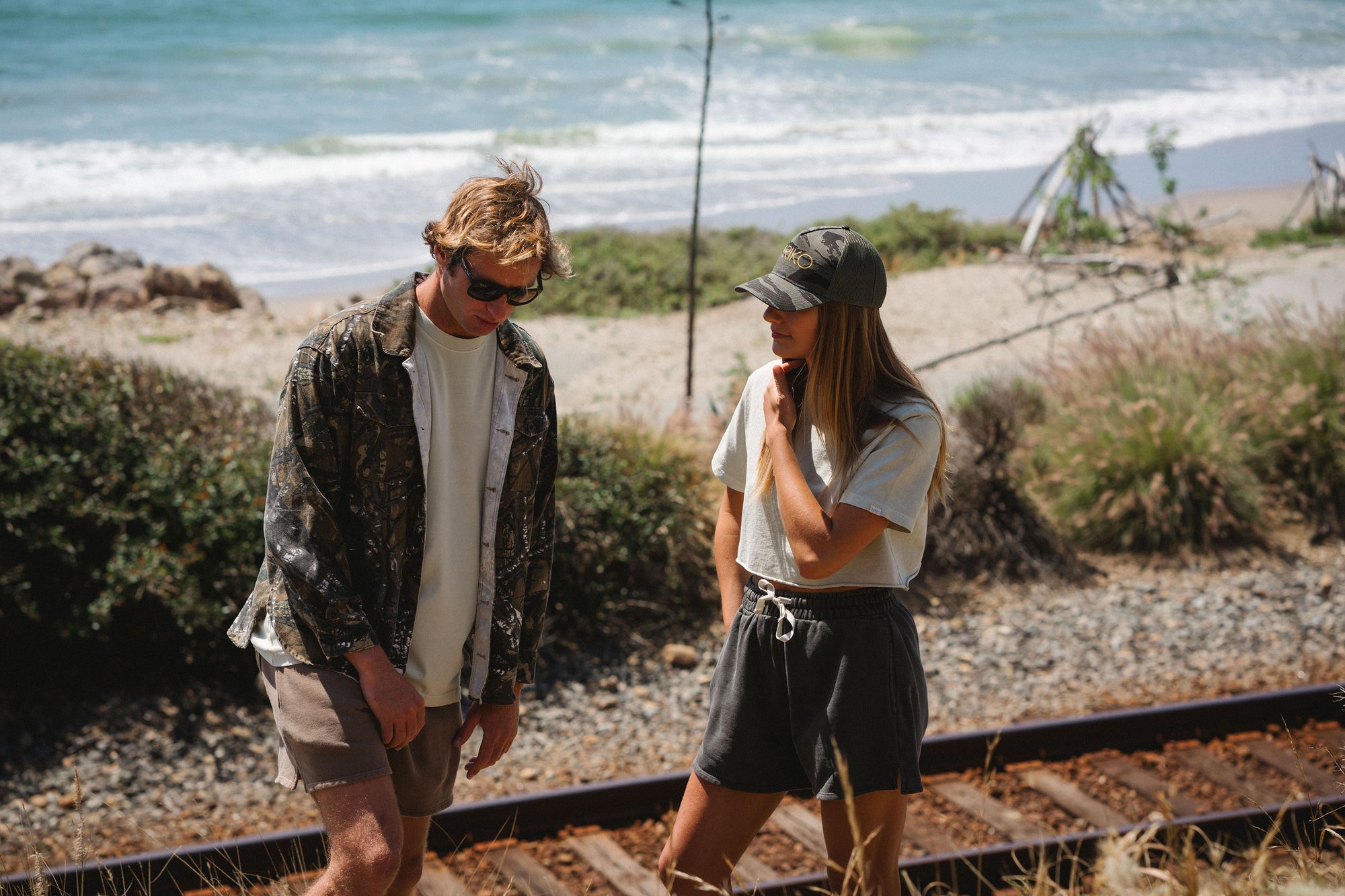 Two people walking along a coastal path with ocean in the background. Man is wearing a real tree camo bucky jacket and brown french terry day shorts. She is wearing a beach crop t-shirt in beige with dark gray french terry day shorts and an ocean tiger camo surf trucker. 