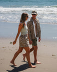Two people walking on a beach with ocean waves in the background. She is wearing a surf tank in beige and leopard camo day shorts. He is wearing a bucky leopard camo jacket, army green day shorts and a leopard camo surf trucker hat. 