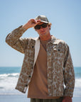 Man wearing a bucky leopard camouflage jacket, brown beach t-shirt, army green day shorts and sunglasses on a beach.