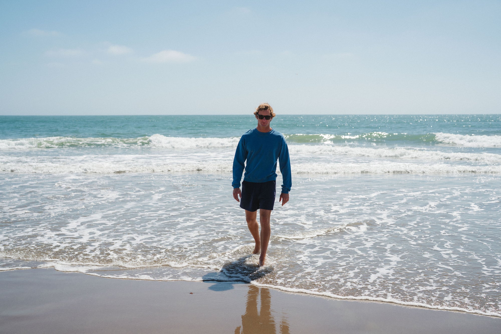 Man walking on a beach with ocean waves in the background. Blue post surf crew sweatshirt and navy day shorts. 