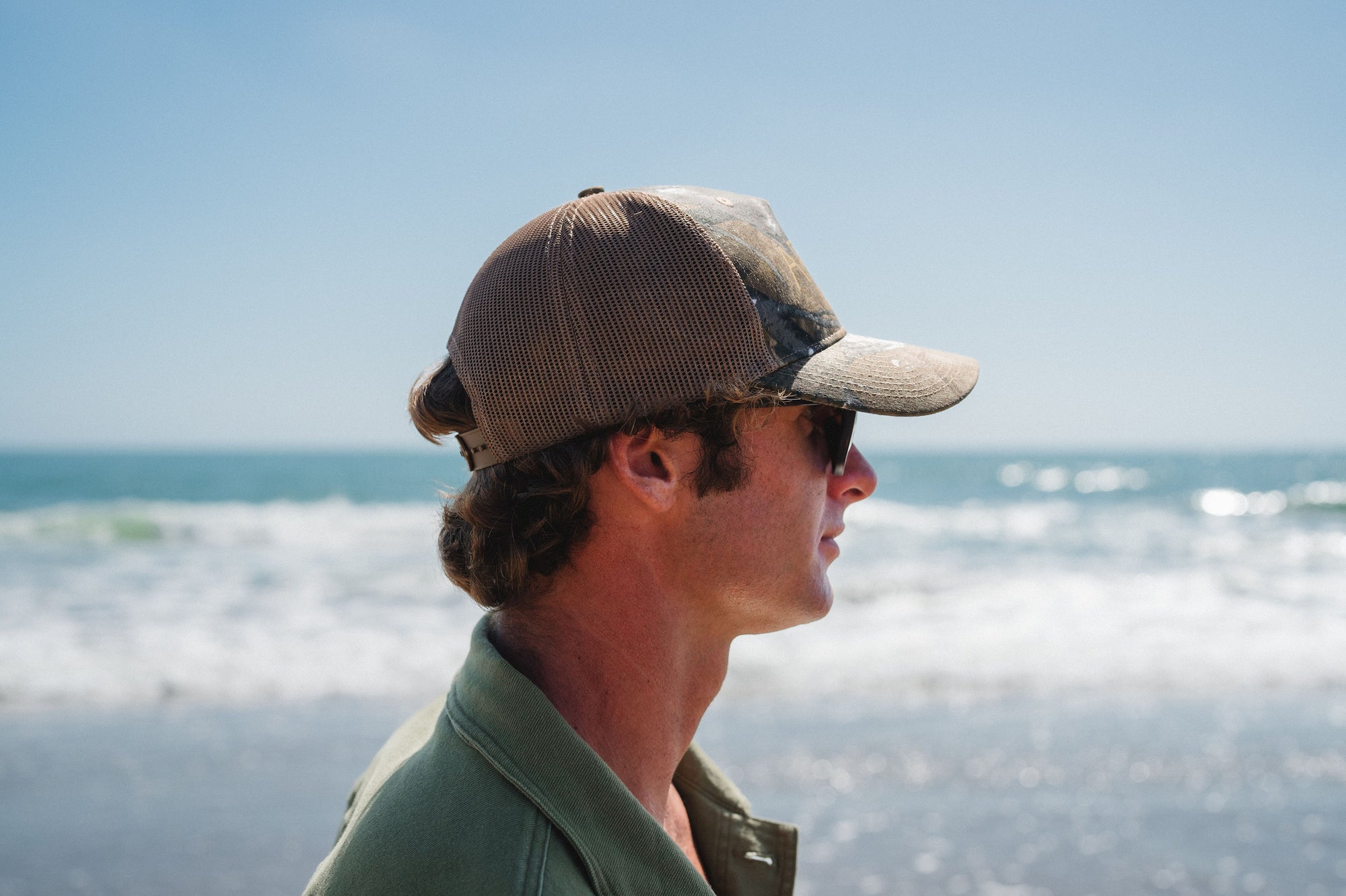 Man wearing a real tree surf trucker hat looking out at the ocean on a beach.