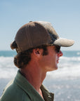 Man wearing a real tree surf trucker hat looking out at the ocean on a beach.