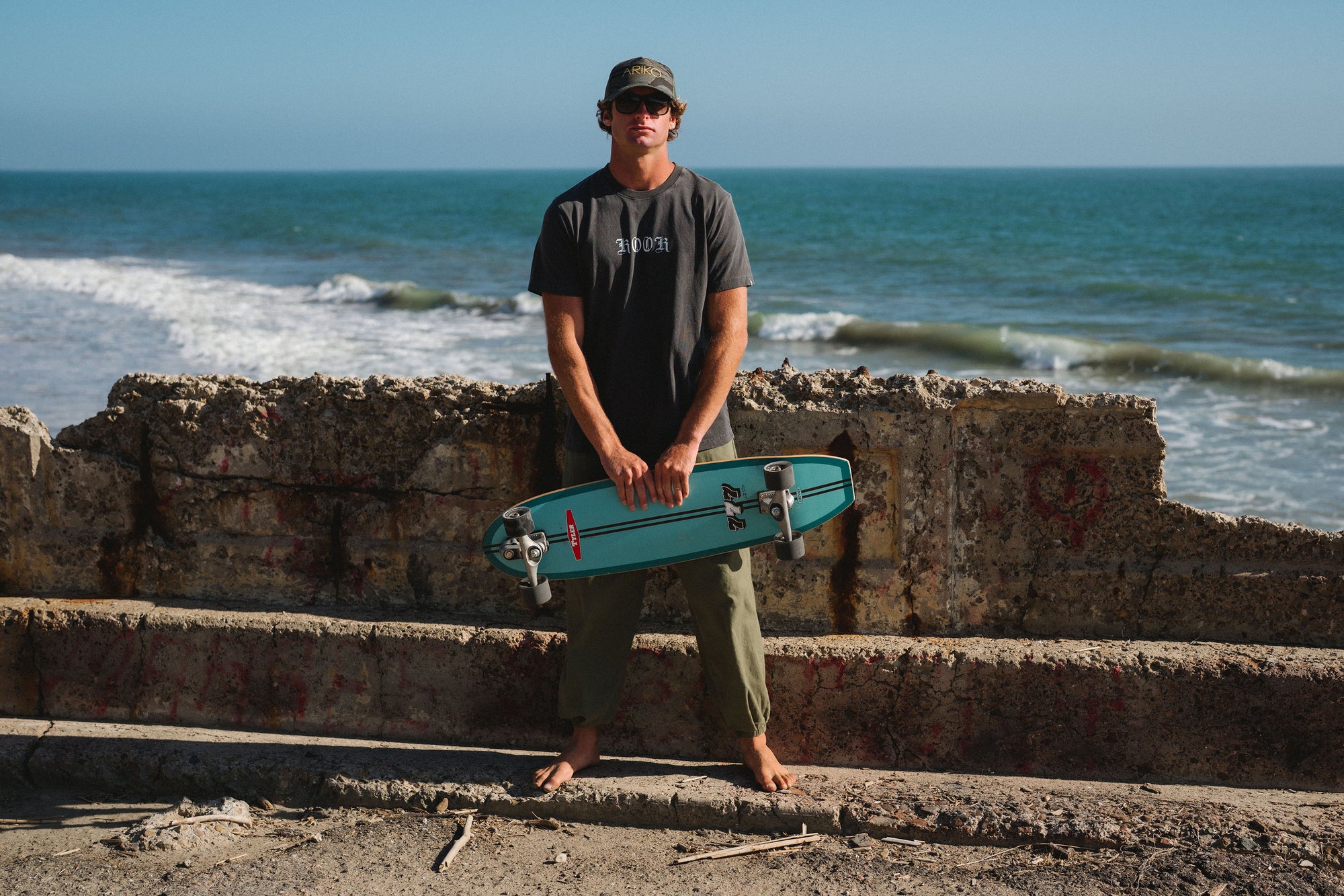Man holding a teal skateboard on a beach with ocean waves in the background. He is wearing a 'KOOK' graphic t-shirt with army green day pants and an ocean tiger surf trucker hat. 