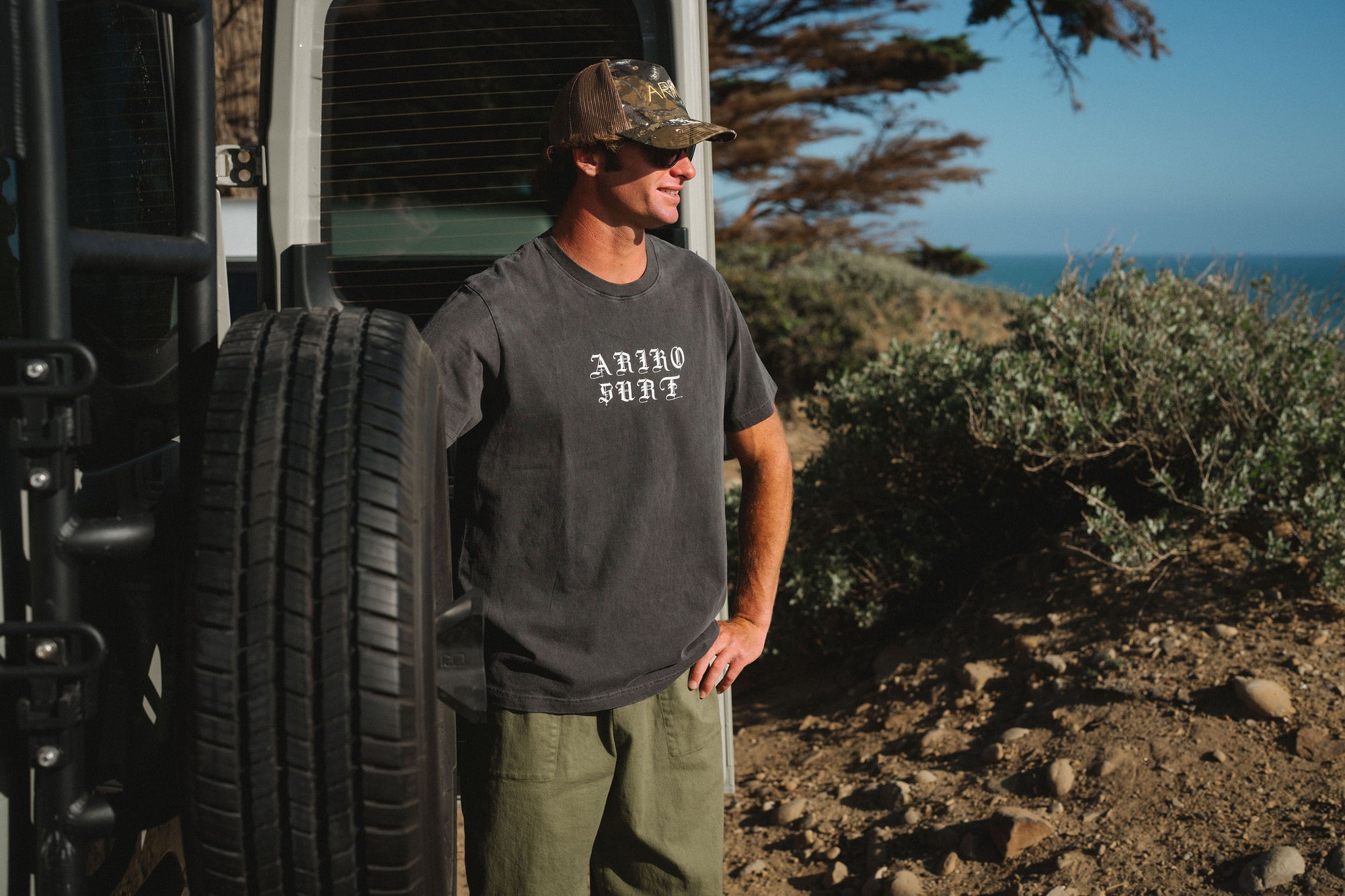 Man standing next to a vehicle with a scenic background. He is wearing a 'ARIKO SURF' graphic t-shirt with army green day pants and a real tree surf trucker hat. 