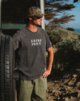 Man standing next to a vehicle with a scenic background. He is wearing a 'ARIKO SURF' graphic t-shirt with army green day pants and a real tree surf trucker hat. 