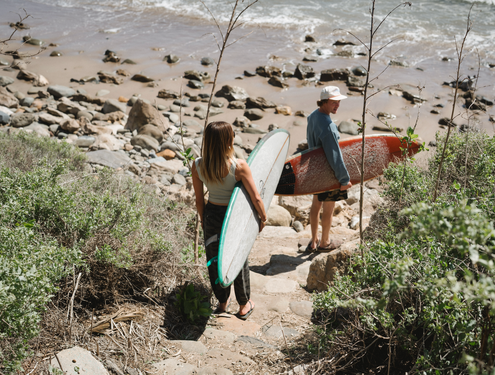 Two people carrying surfboards walking up a rocky path towards the ocean. Wearing ARIKO clothing.