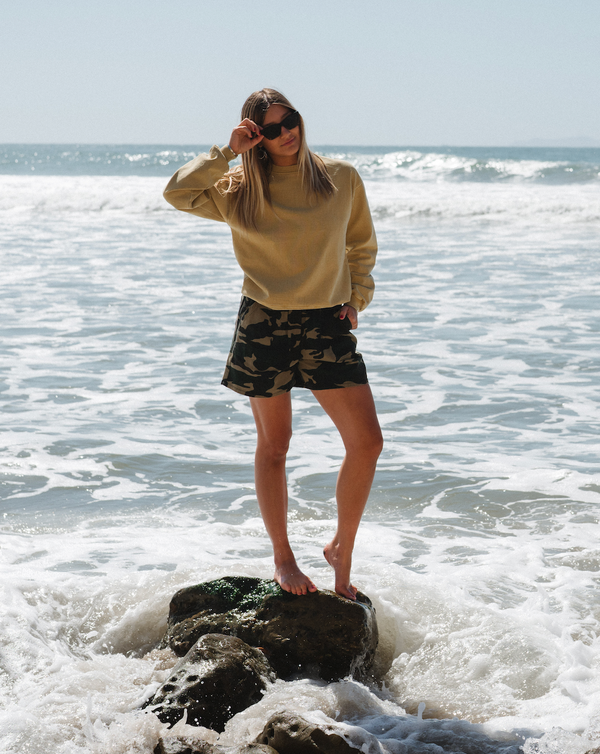 woman on a rock in shallow ocean wearing post surf crew sweatshirt in sunrise yellow with camo day shorts