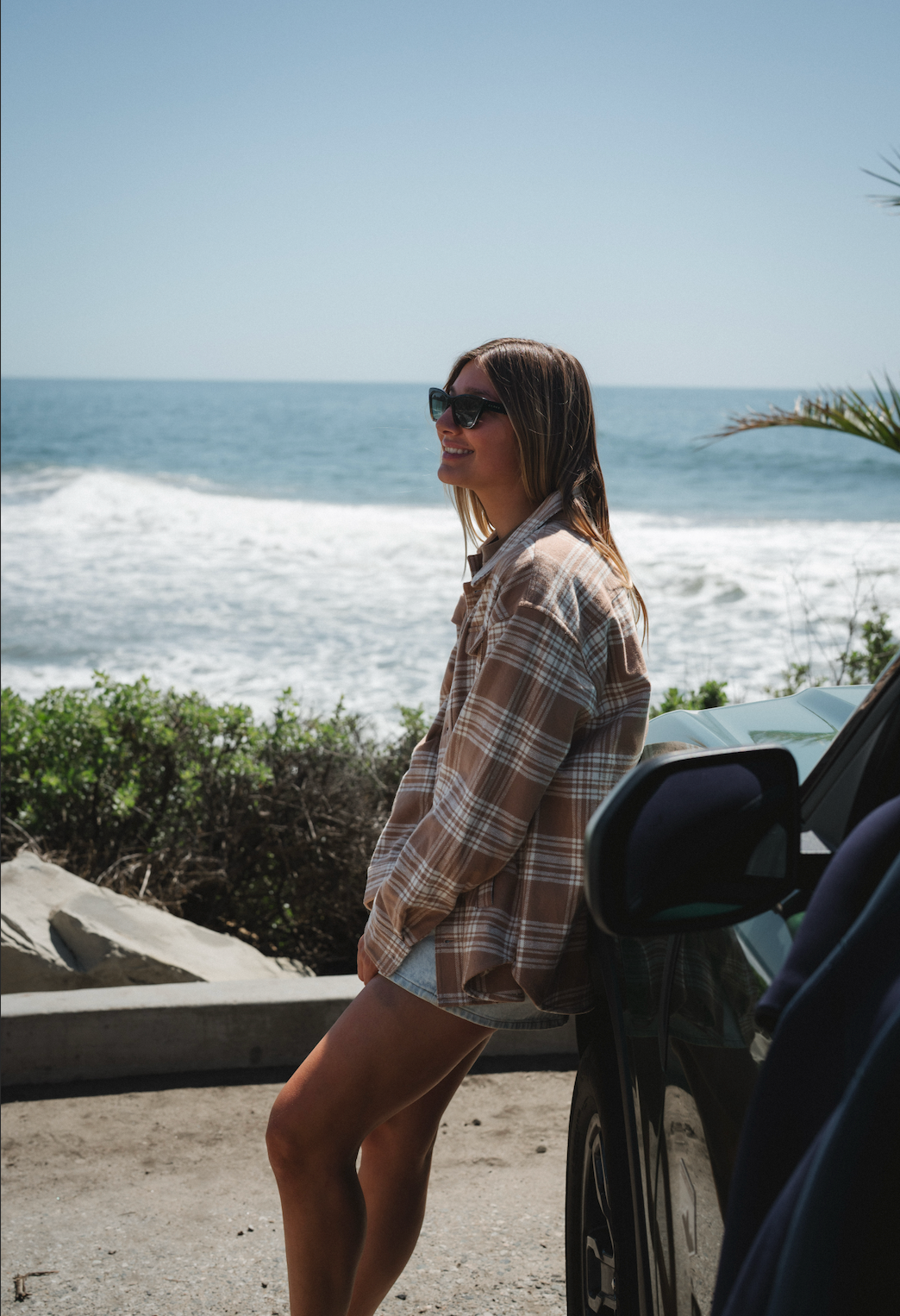 Woman standing by a car near the ocean with sunglasses on wearing Bucky Split Hem Shirt Jacket in Beige/Plaid