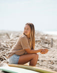 Woman sitting on a beach wearing the beach t-shirt in brownstone. 